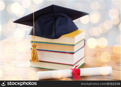 education, school, graduation and knowledge concept - close up of books and mortarboard with diploma on wooden table over holidays lights background
