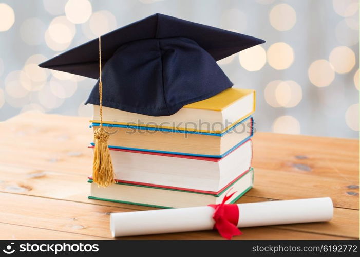 education, school, graduation and knowledge concept - close up of books and mortarboard with diploma on wooden table over holidays lights background