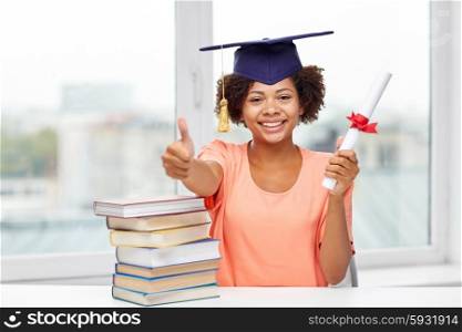 education, school, graduation, gesture and people concept - happy smiling african american student girl in bachelor cap with books and diploma sitting at table and showing thumbs up at home