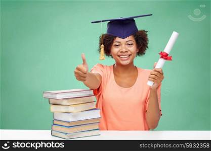 education, school, graduation, gesture and people concept - happy smiling african american student girl in bachelor cap with books and diploma showing thumbs up over green chalk board background