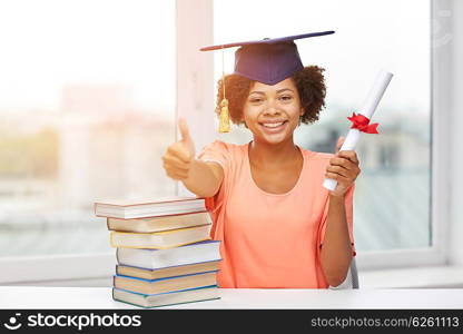 education, school, graduation, gesture and people concept - happy smiling african american student girl in bachelor cap with books and diploma sitting at table and showing thumbs up at home