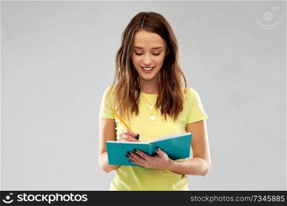 education, school, inspiration and people concept - smiling young woman or teenage student girl in yellow t-shirt writing to diary or notebook by pencil over grey background. teenage student girl writing to diary or notebook