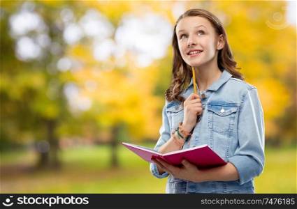 education, school, inspiration and people concept - teenage student girl in denim jacket with diary or notebook and pencil thinking over autumn park background. teenage student girl with diary or notebook