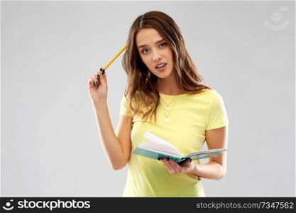 education, school, inspiration and people concept - young woman or teenage student girl in yellow t-shirt with diary or notebook and pencil thinking hard over grey background. teenage student girl with diary or notebook thinks