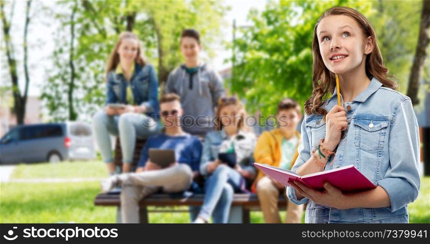 education, school, inspiration and people concept - young woman or teenage student girl in denim jacket with diary or notebook and pencil thinking over group of friends outdoors background. teenage student girl with diary or notebook