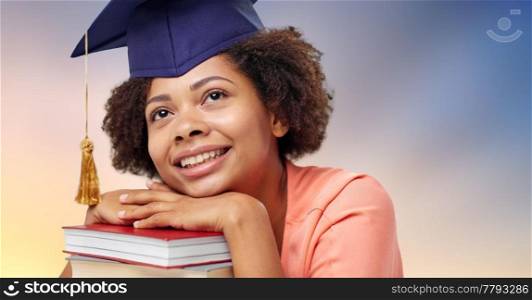 education, school, knowledge and graduation concept - close up of happy smiling african american graduate student girl in mortarboard with books dreaming. close up of african graduate student with books