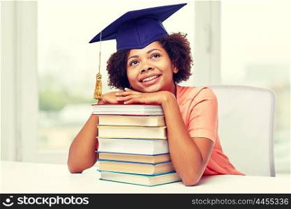 education, school, knowledge and people concept - happy smiling african american student girl in bachelor cap with books sitting at table and dreaming at home