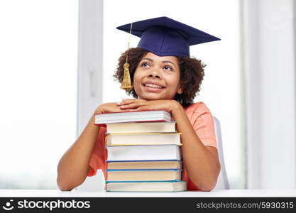 education, school, knowledge and people concept - happy smiling african american student girl in bachelor cap with books sitting at table and dreaming at home