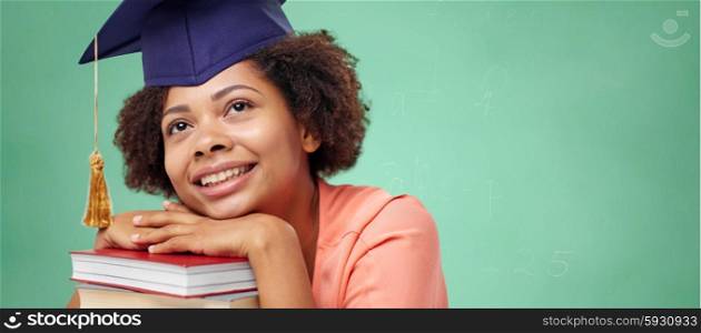 education, school, knowledge and people concept - happy smiling african american student girl in bachelor cap with books sitting at table and dreaming over green chalk board background