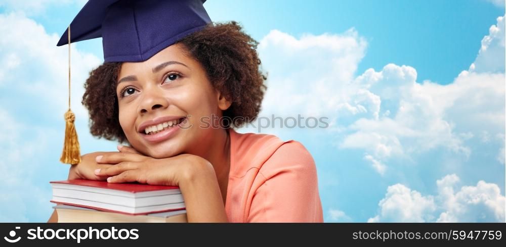 education, school, knowledge and people concept - happy smiling african american student girl in bachelor cap with books sitting at table and dreaming over blue sky and clouds background