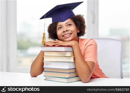 education, school, knowledge and people concept - happy smiling african american student girl in bachelor cap with books sitting at table and dreaming at home