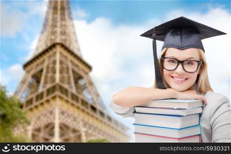 education, school, knowledge and people concept - picture of happy student girl or woman in trencher cap with stack of books over paris eiffel tower background