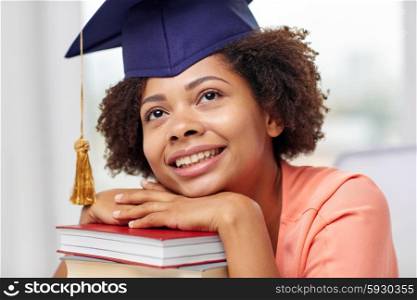 education, school, knowledge, graduation and people concept - happy smiling african american student girl in bachelor cap with books sitting at table and dreaming at home