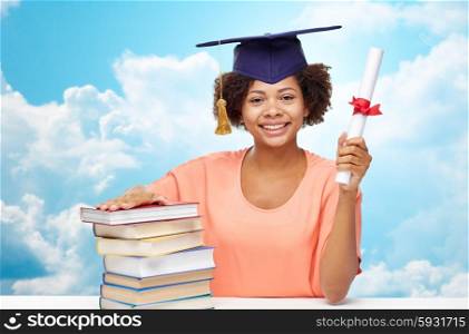 education, school, knowledge, graduation and people concept - happy smiling african american student girl in bachelor cap with books and diploma sitting at table over blue sky and clouds background
