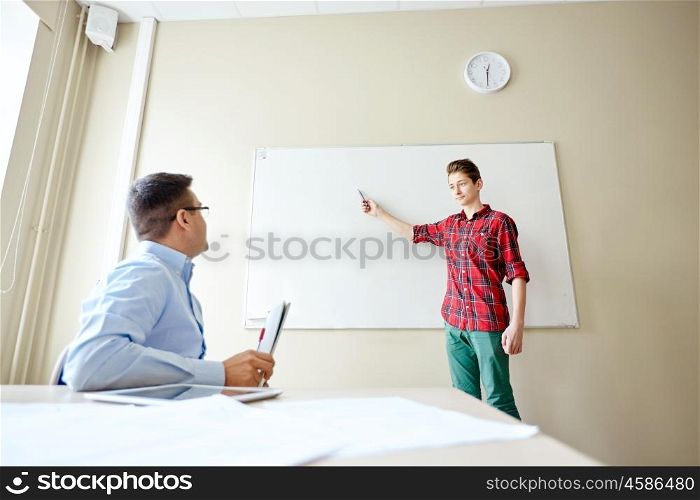 education, school, learning and people concept - student boy showing something on blank white board and teacher in classroom