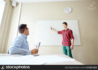 education, school, learning and people concept - student boy showing something on blank white board and teacher in classroom