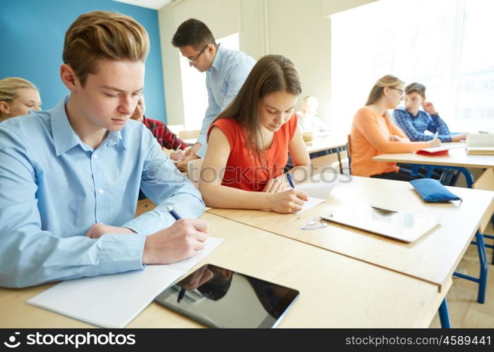education, school, learning, teaching and people concept - group of students writing test or exam and teacher in classroom