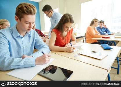 education, school, learning, teaching and people concept - group of students writing test or exam and teacher in classroom