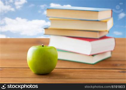 education, school, literature, reading and knowledge concept - close up of books and green apple on wooden table over blue sky and clouds background
