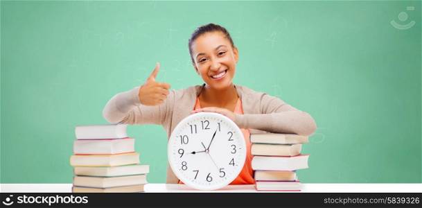 education, school, people and learning concept - afro american student girl with books and clock showing thumbs up over green chalk board background