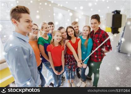 education, school, technology and people concept - group of happy smiling students taking picture with smartphone selfie stick in corridor over snow. group of students taking selfie with smartphone