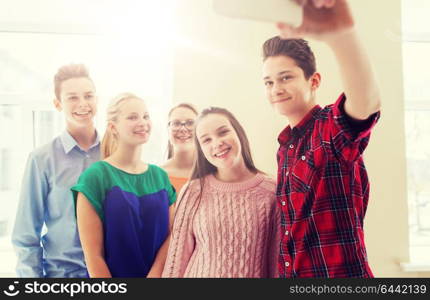 education, school, technology and people concept - group of happy smiling students taking picture with smartphone selfie stick in corridor. group of students taking selfie with smartphone