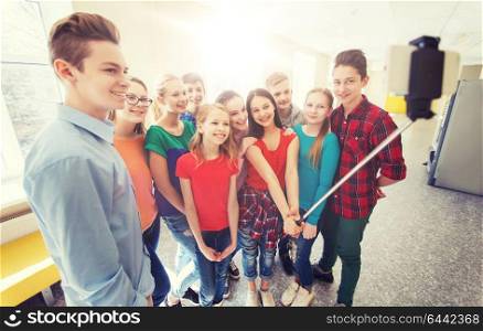 education, school, technology and people concept - group of happy smiling students taking picture with smartphone selfie stick in corridor. group of students taking selfie with smartphone