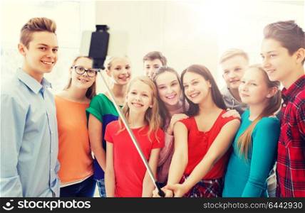 education, school, technology and people concept - group of happy smiling students taking picture with smartphone selfie stick in corridor. group of students taking selfie with smartphone