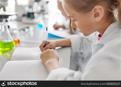 education, science and children concept - close up of girl studying chemistry at school laboratory and writing to workbook. girl studying chemistry at school laboratory