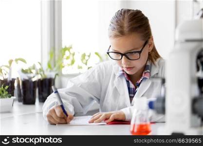 education, science and children concept - girl studying chemistry at school laboratory and writing to workbook. girl studying chemistry at school laboratory