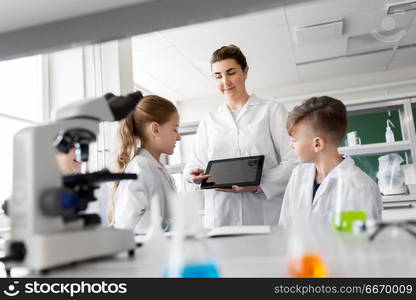 education, science and technology concept - chemistry teacher showing tablet pc computer to kids or students at school laboratory. teacher with tablet pc and kids at chemistry class. teacher with tablet pc and kids at chemistry class