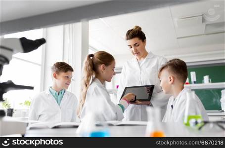education, science and technology concept - chemistry teacher showing tablet pc computer to kids or students at school laboratory. teacher with tablet pc and kids at chemistry class. teacher with tablet pc and kids at chemistry class