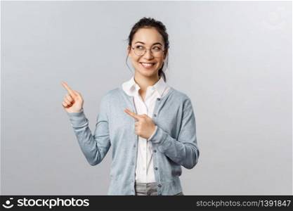 Education, studying and online classes concept. Intrigued, excited happy asian woman in glasses peeking left, smiling and pointing upper corner at advertisement or chart, grey background.. Education, studying and online classes concept. Intrigued, excited happy asian woman in glasses peeking left, smiling and pointing upper corner at advertisement or chart, grey background