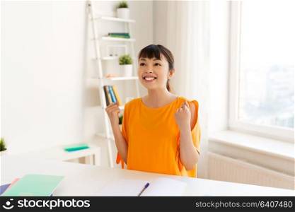 education, success, learning and people concept - happy smiling asian student girl with notebooks making fist pump gesture at home. asian student girl celebrating success at home