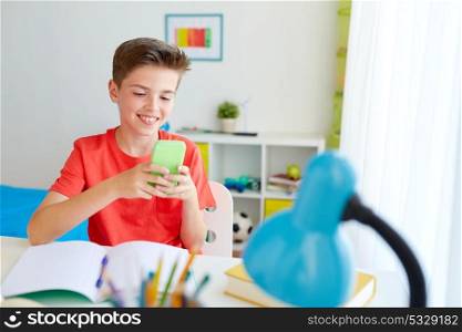 education, technology and communication concept - happy student boy distracting from homework and texting on smartphone at home. student boy with smartphone distracting from study