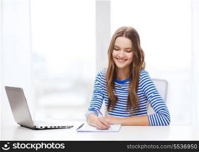 education, technology and home concept - smiling teenage girl with laptop computer, notebook and pen at home