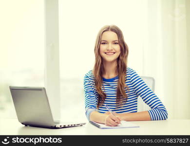 education, technology and home concept - smiling teenage girl with laptop computer, notebook and pen at home