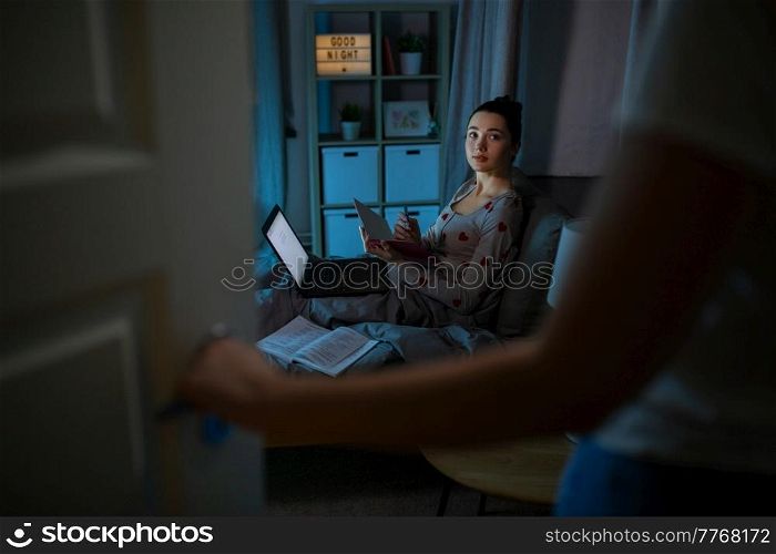 education, technology and people concept - teenage student girl with notebook and laptop computer learning in bed at home at night and mother entering room. student girl with laptop learning at night