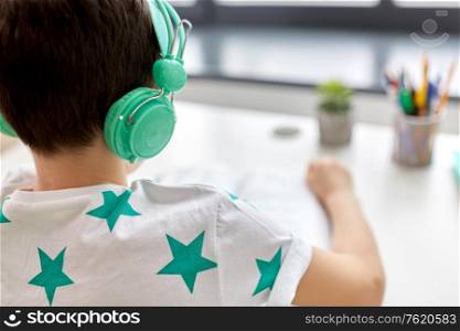 education, technology and school concept - close up of boy in headphones sitting at table at home. boy in headphones with textbook learning at home