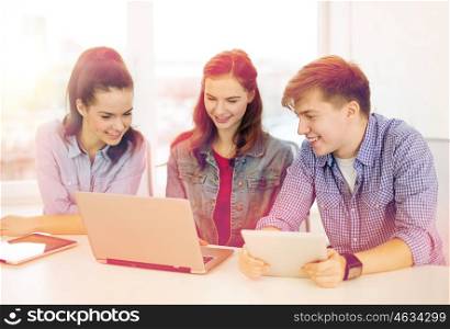 education, technology, school and internet concept - three smiling students with laptop and tablet pc at school