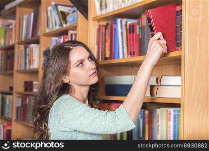 Education theme image with a beautiful girl taking a book from a library in order to study or enjoying a novel in the spare time, dressed in a cozy outfit.