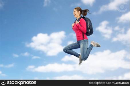 education, travel, tourism, motion and people concept - smiling young woman or student with backpack jumping in air over blue sky and clouds background