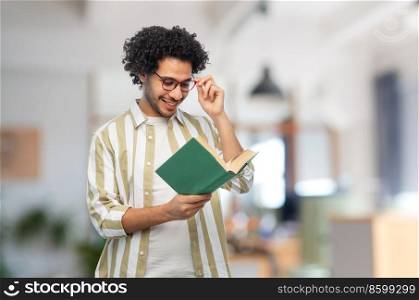 education, vision and people concept - happy smiling young man in glasses reading book over office background. happy young man in glasses reading book at office