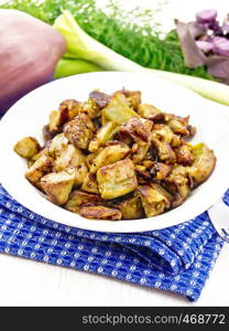 Eggplant fried with onions, eggs and spices in a plate on a blue napkin, basil, leek and dill on wooden board background