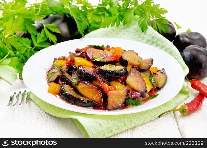 Eggplant fried with plums, onions and sweet peppers in a plate on a towel against the background of wooden board