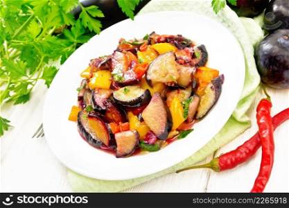 Eggplant fried with plums, onions and sweet peppers in a plate on a napkin against light wooden board