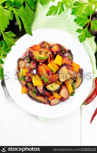 Eggplant fried with plums, onions and sweet peppers in a plate on a towel against background of wooden board from above