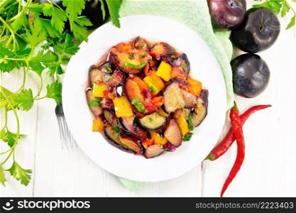 Eggplants fried with plums, onions and sweet peppers in a plate on a napkin on background of wooden board from above