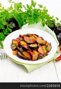 Eggplants fried with plums, onions and sweet peppers in a plate on a towel against the background of white wooden board