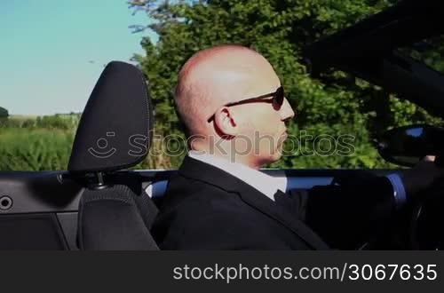 ein Mann in einem schwarzen Business-Anzug fahrt in einem Cabrio durch eine grune Landschaft